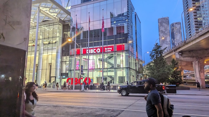 A modern urban scene featuring a glass-fronted building with a sign reading 'CIBC Banking Centre.' The setting is in a city environment with skyscrapers visible in the background. There's a road in front with pedestrians walking and a pickup truck driving by. It's evening or early night, as indicated by the lighting and the sky's color.