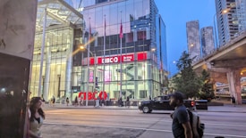 A modern urban scene featuring a glass-fronted building with a sign reading 'CIBC Banking Centre.' The setting is in a city environment with skyscrapers visible in the background. There's a road in front with pedestrians walking and a pickup truck driving by. It's evening or early night, as indicated by the lighting and the sky's color.