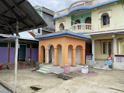 A colorful residential building with intricate architectural details is shown in the foreground. The building has a pastel yellow facade with arched windows and balcony railings. A separate smaller structure with orange columns is situated in the courtyard, surrounded by an open area with concrete flooring. Two women are seated in the courtyard, one on a step and another near the smaller structure. The setting has a casual and relaxed atmosphere.