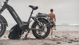 A large, rugged-looking e-bike parked on a sandy beach with a shirtless person walking towards the ocean. The bike has a sturdy frame and oversized tires, suitable for off-road terrain. The sky is overcast, creating a calm and subdued atmosphere.