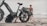 A large, rugged-looking e-bike parked on a sandy beach with a shirtless person walking towards the ocean. The bike has a sturdy frame and oversized tires, suitable for off-road terrain. The sky is overcast, creating a calm and subdued atmosphere.