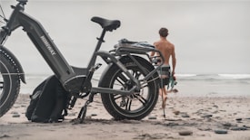 A large, rugged-looking e-bike parked on a sandy beach with a shirtless person walking towards the ocean. The bike has a sturdy frame and oversized tires, suitable for off-road terrain. The sky is overcast, creating a calm and subdued atmosphere.