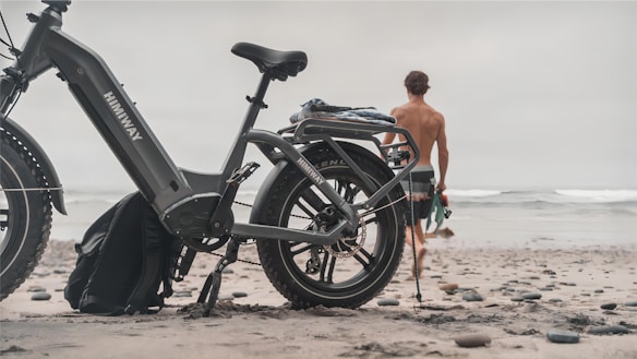 A large, rugged-looking e-bike parked on a sandy beach with a shirtless person walking towards the ocean. The bike has a sturdy frame and oversized tires, suitable for off-road terrain. The sky is overcast, creating a calm and subdued atmosphere.