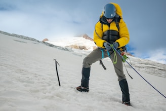 A climber wearing a yellow jacket and helmet is securely adjusting ropes on a snowy slope. An ice axe is embedded in the snow nearby. The background shows a mountainous landscape under a partly cloudy sky.