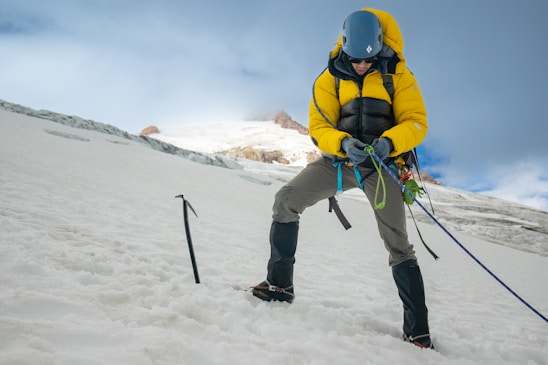 A climber wearing a yellow jacket and helmet is securely adjusting ropes on a snowy slope. An ice axe is embedded in the snow nearby. The background shows a mountainous landscape under a partly cloudy sky.