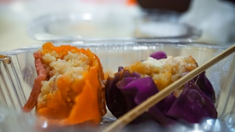 Two pieces of brightly colored dim sum rest inside a transparent plastic container. One dumpling is wrapped in an orange skin, while the other has a purple exterior. A chopstick lies across the container.