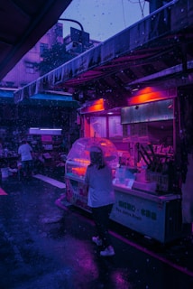 A street market scene at night featuring a brightly lit food stall with glowing pink and purple neon lights. The area is wet, suggesting recent rainfall, with a person under an umbrella walking past. The surrounding environment includes additional stalls and urban buildings, dimly lit by the ambient streetlights.