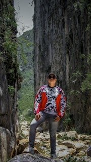 A person dressed in a colorful, patterned jacket and sunglasses stands confidently on a rocky path surrounded by high, green-covered cliffs. The scene is rich with greenery and rugged natural elements, evoking a sense of adventure.