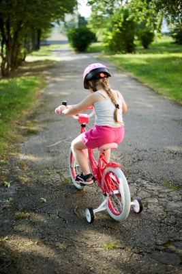 A young child with braided hair rides a red bicycle with training wheels along a paved path surrounded by green grass and trees. The child is wearing a pink helmet, pink shorts, and black sandals.