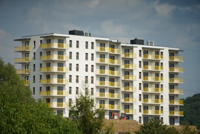 A modern, multi-story residential building with white walls and striking yellow balconies is set against a partly cloudy sky. Trees and vegetation are visible in the foreground, and there is a piece of construction equipment near the building.