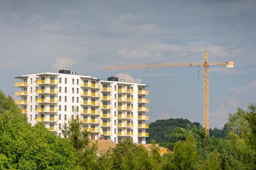 A modern multi-story residential building with white walls and yellow balconies is located near a crane. The structure is surrounded by lush green trees and set against a partly cloudy sky.