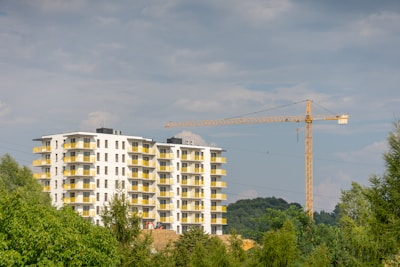 A modern apartment building with multiple floors and yellow balcony railings, surrounded by greenery. In the background, a tall construction crane is visible, indicating ongoing construction work. The sky is partly cloudy.