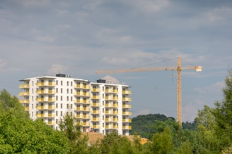 A modern apartment building with multiple floors and yellow balcony railings, surrounded by greenery. In the background, a tall construction crane is visible, indicating ongoing construction work. The sky is partly cloudy.