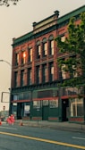 An older brick building with green accents is featured prominently. The words 'Hull Building' are visible at the top. The ground floor windows are boarded up, with a 'For Lease' sign in one window. On the sidewalk, a construction sign and traffic cone are present. A tree partially obscures the right side of the building.