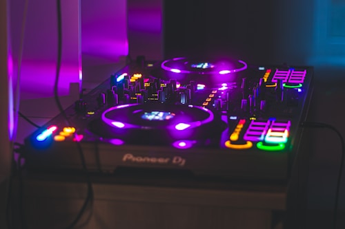 A DJ controller with illuminated lights and buttons in various colors sits on a surface. The scene is bathed in purple lighting, creating a vibrant and energetic atmosphere.