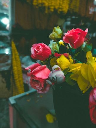 A vibrant bouquet of pink and yellow flowers, prominently featuring roses, placed in a dark vase. The background appears to be a market setting with yellow marigold garlands hanging, creating a lively yet slightly blurred atmosphere.