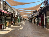 A narrow, cobblestone street lined with cozy shops on both sides. The street is covered with fabric shade sails, creating an artistic pattern. Several potted plants and classic street lamps add charm to the setting, with a bright blue sky visible above.