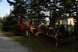 Two heavy machinery vehicles are situated on a grassy area near a forest. One is a red tractor, and the other is a green bulldozer. They are positioned under the shade of tall pine trees, with a dirt path visible beside them.