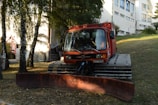 A red snow groomer with a large plow blade attached at the front is parked on a grassy area near a building complex. Several trees are present in the background, casting shadows on the ground. The building in the background appears to be multi-storied with multiple windows.