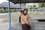 A person in a light brown scout uniform and dark brown hijab is sitting on a wooden platform under a metal roof. There is a school building in the background with blue and beige walls, and neatly trimmed bushes in front. The area is open and grassy, with trees and additional structures visible further back.