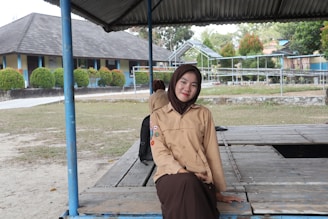 A person in a light brown scout uniform and dark brown hijab is sitting on a wooden platform under a metal roof. There is a school building in the background with blue and beige walls, and neatly trimmed bushes in front. The area is open and grassy, with trees and additional structures visible further back.