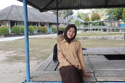 A person in a light brown scout uniform and dark brown hijab is sitting on a wooden platform under a metal roof. There is a school building in the background with blue and beige walls, and neatly trimmed bushes in front. The area is open and grassy, with trees and additional structures visible further back.