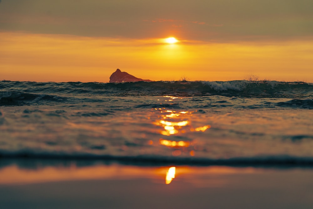 the sun is setting over the ocean with a rock in the distance