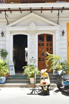 A quaint entrance to a building features a decorative pediment and wooden door, flanked by lush green plants in ceramic pots. A yellow scooter and a blue motorcycle are parked in front, while a sign indicates that bookings are open.