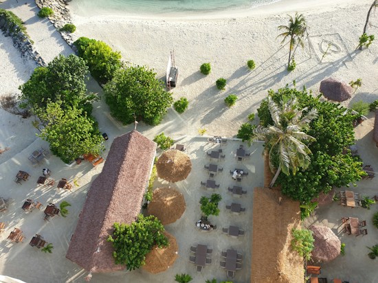 An aerial view of a beachfront area featuring lush green trees, sandy pathways, and organized dining setups with wooden tables and chairs. A thatched-roof structure is centrally located, surrounded by palm trees and green foliage. There are two larger thatched umbrellas providing shade. The beach stretches towards the calm turquoise sea.