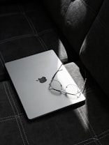 A pair of eyeglasses rests on a closed laptop with a visible logo, placed on a dark, textured couch. Sunlight casts a narrow beam, creating a gentle contrast with the shadows.