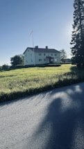 A large, two-story white house with rectangular windows stands prominently in an open grassy field. A flagpole with a small flag is situated next to the house, and tall trees border the property. The sun casts long shadows across the grass and nearby road, suggesting early morning or late afternoon.