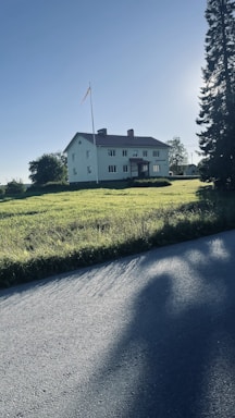 A large, two-story white house with rectangular windows stands prominently in an open grassy field. A flagpole with a small flag is situated next to the house, and tall trees border the property. The sun casts long shadows across the grass and nearby road, suggesting early morning or late afternoon.