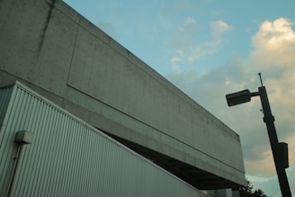 A concrete building features a textured facade with a modern, industrial design. A streetlight is visible in the foreground, and the sky is partly cloudy with hints of blue and orange tones.