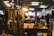 A collection of vintage and cultural items displayed on a wooden shelf, including various metal necklaces, ornamental beads, and small statues of Buddha. The scene is warmly lit by overhead lamps, creating a cozy and inviting atmosphere. In the background, there is a soft focus of a blurred indoor market setting.