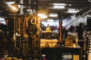 A collection of vintage and cultural items displayed on a wooden shelf, including various metal necklaces, ornamental beads, and small statues of Buddha. The scene is warmly lit by overhead lamps, creating a cozy and inviting atmosphere. In the background, there is a soft focus of a blurred indoor market setting.