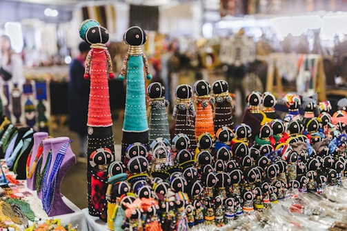 A collection of intricately crafted beaded dolls is arranged on a table at a market. The dolls vary in size and are adorned with brightly colored beads and fabric, each featuring black heads with simple faces. In the background, various other handmade crafts and necklaces are visible, adding to the vibrant and lively atmosphere of the market setting.