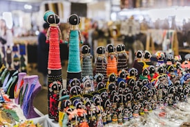 A collection of intricately crafted beaded dolls is arranged on a table at a market. The dolls vary in size and are adorned with brightly colored beads and fabric, each featuring black heads with simple faces. In the background, various other handmade crafts and necklaces are visible, adding to the vibrant and lively atmosphere of the market setting.