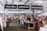 An indoor market scene features several stalls displaying various items, including clothing, crafts, and food. Signs above direct visitors to areas such as a chill zone, rooftop bar, and deli. People are browsing, interacting, and shopping, while mannequins and displays add color to the setting.