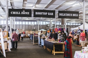 An indoor market scene features several stalls displaying various items, including clothing, crafts, and food. Signs above direct visitors to areas such as a chill zone, rooftop bar, and deli. People are browsing, interacting, and shopping, while mannequins and displays add color to the setting.