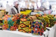 Vibrant, colorful bags with African-inspired geometric patterns are displayed on a table in a market setting. A person wearing a fur coat is blurred in the background, suggesting a busy outdoor market atmosphere.