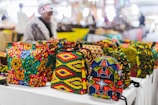 Vibrant, colorful bags with African-inspired geometric patterns are displayed on a table in a market setting. A person wearing a fur coat is blurred in the background, suggesting a busy outdoor market atmosphere.