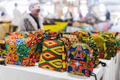 Vibrant, colorful bags with African-inspired geometric patterns are displayed on a table in a market setting. A person wearing a fur coat is blurred in the background, suggesting a busy outdoor market atmosphere.