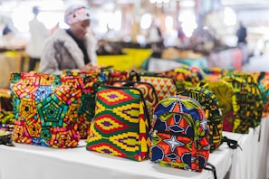 Vibrant, colorful bags with African-inspired geometric patterns are displayed on a table in a market setting. A person wearing a fur coat is blurred in the background, suggesting a busy outdoor market atmosphere.