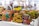 Vibrant, colorful bags with African-inspired geometric patterns are displayed on a table in a market setting. A person wearing a fur coat is blurred in the background, suggesting a busy outdoor market atmosphere.