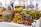 Vibrant, colorful bags with African-inspired geometric patterns are displayed on a table in a market setting. A person wearing a fur coat is blurred in the background, suggesting a busy outdoor market atmosphere.