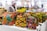 Vibrant, colorful bags with African-inspired geometric patterns are displayed on a table in a market setting. A person wearing a fur coat is blurred in the background, suggesting a busy outdoor market atmosphere.