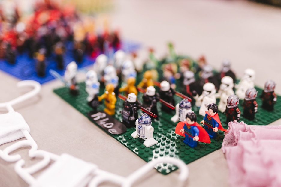 A collection of colorful LEGO minifigures is arranged on a green baseplate. The figures include a variety of characters, some with capes and helmets, and others representing iconic figures. In the background, more figures are placed on a blue baseplate. Nearby are white hangers and a piece of fabric.