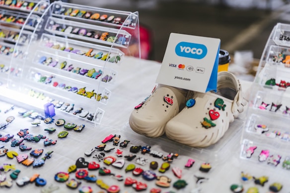 A table displaying a collection of decorative shoe charms and two beige clogs adorned with colorful designs, including a mushroom and heart. The background features clear stands holding additional charms. A sign with 'YOCO' branding and payment options is present.
