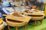 Bowls filled with delicious-looking stir-fried noodles, garnished with fresh lime and vegetables, are placed on woven bamboo stands. Wooden boards serve as trays supporting the bowls, suggesting an inviting and rustic presentation. The blurred background hints at a busy market or food stall atmosphere.