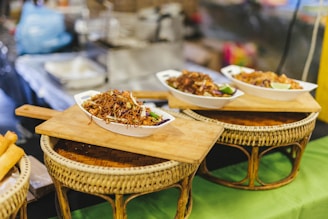 Bowls filled with delicious-looking stir-fried noodles, garnished with fresh lime and vegetables, are placed on woven bamboo stands. Wooden boards serve as trays supporting the bowls, suggesting an inviting and rustic presentation. The blurred background hints at a busy market or food stall atmosphere.